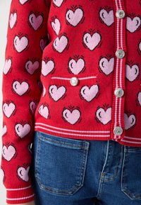 Punainen neulepusero, jossa on vaaleanpunaisia sydänkuvioita, joissa on hopeiset napit ja raidallinen vyötärö. Yhdistetty sinisiin farkkuihin.