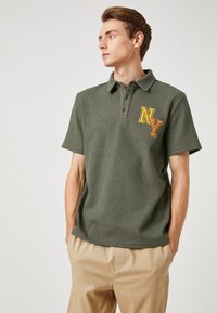 Oliwkowa koszulka polo z krótkim rękawem, ożebrowanym wzorze i żółtym haftowanym logo "NY" na lewej piersi. Klasyczny design kołnierza.
