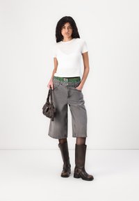 T-shirt bianco a maniche corte, pantaloni di denim grigi a gamba larga con cintura a quadri verde, e stivali al ginocchio marroni scuri. Borsa marrone in mano.
