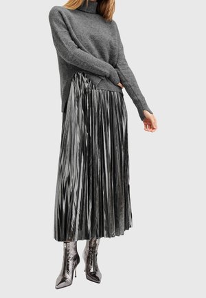 Kobieta ubrana w szary sweter z golfem, metaliczną plisowaną spódnicę midi oraz pasujące botki na obcasie z ozdobnymi zamkami z przodu.