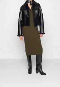 Svart läderjacka i kort modell med fårskinnskrage, olivgrön ribbad midiklänning och knähöga svarta stövlar med klack. Sjal knuten runt halsen.