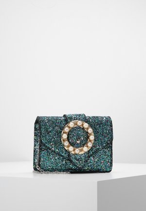 Mała błyszcząca torebka w kolorze morskim z okrągłą złotą klamrą zdobioną perłami i srebrnym łańcuszkiem na ramię, prezentowana na białym cokole.