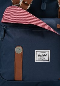 Granatowy plecak z materiału z brązowymi skórzanymi akcentami, przednia kieszeń na zamek błyskawiczny, w paski czerwone wnętrze i metka marki Herschel. Teksturowany materiał.