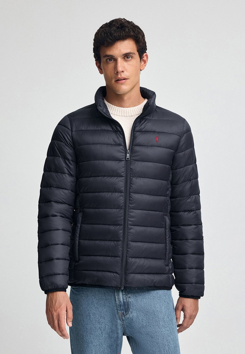 Granatowa kurtka puchowa z stójką, zamek błyskawiczny i boczne kieszenie. Wykonana z lekkiego materiału, z pikowaną fakturą i czerwoną logotypem.