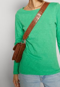 Camicia verde a maniche lunghe abbinata a una borsa a tracolla in pelle marrone con texture, dotata di una tracolla decorata e dettagli in metallo.