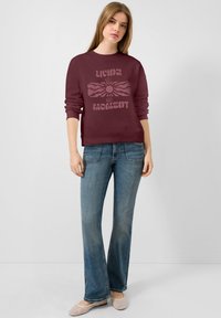 Sweatshirt bordeaux avec le texte graphique "Vivre l'instant présent", associé à un jean bleu évasé et des ballerines claires. Tissu lisse, coupe décontractée.
