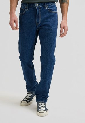 Mann trägt dunkelblaue Jeans und schwarze Turnschuhe mit weißen Schnürsenkeln, steht vor einem schlichten hellen Hintergrund.
