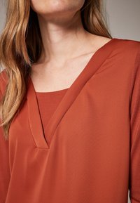 Top color ruggine con design a V, look a strati con un collo interno quadrato e una texture di tessuto morbida e liscia.