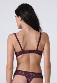 Reggiseno in pizzo color mogano con spalline regolabili e chiusura a gancio. Presenta un motivo floreale e bordi scallop. Perizoma in pizzo color mogano coordinato.