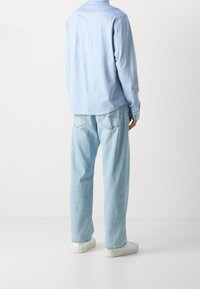 Hellblaues Langarmhemd mit lockerer Passform und mit Knopfmanschetten, kombiniert mit hellblauen Jeans mit geradem Bein und weißen Turnschuhen. Schlichtes Design.