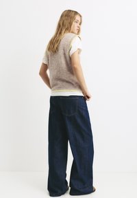 Beige Strickweste mit hellgelben Akzenten, getragen über einem weißen T-Shirt, kombiniert mit lockeren dunkelblauen Jeans. Weiche Textur und bequeme Passform.