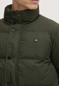 Green puffer jacket with a high collar, front snap closures, and small logo patch. Material appears puffy, with a smooth texture.