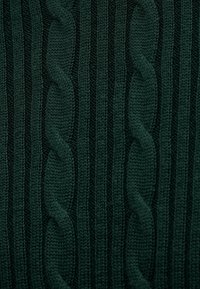 Σκούρο πράσινο πλεκτό ύφασμα που διαθέτει ανυψωμένα σχέδια καλωδίων και κατακόρυφες ραβδώσεις, επιδεικνύοντας υφή και περίπλοκες λεπτομέρειες σε όλη την επιφάνεια.