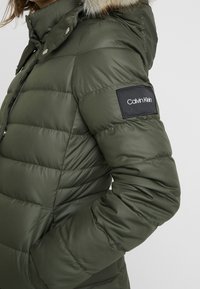 Doudoune vert olive avec capuche amovible doublée de fourrure, fermeture éclair, design matelass é et patch logo Calvin Klein noir sur la manche.