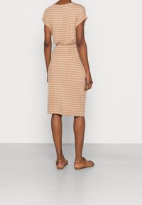 Frau steht, trägt ein beige-weiß gestreiftes knielanges Kleid mit kurzen Ärmeln und braunen flachen Sandalen.