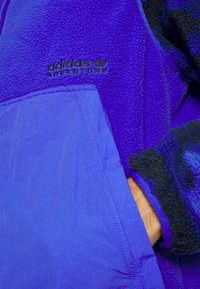 Chaqueta de forro polar morada con un tejido texturizado, un bolsillo suave morado en contraste y un logo "adidas Adventure" en negro.