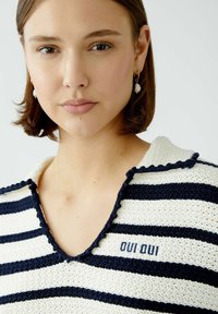 Gestreifter Strickpullover in Creme und Marineblau, mit V-Ausschnitt und gewelltem Kragen, und besticktem Text "OUI OUI" auf der Brust.