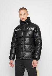 Črna puffer jakna iz umetnega usnja, z visokim ovratnikom, sprednjim zadrgo in belimi logotipnimi poudarki na prsih.