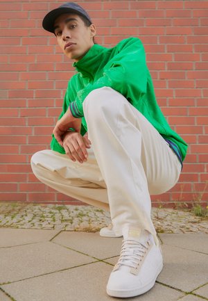 Jeune personne portant une veste verte, un pantalon blanc, des baskets blanches et une casquette sombre accroupie contre un mur en briques rouges.
