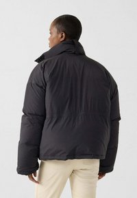 Zwarte pufferjas met een hoge kraag, voorzien van een ontspannen pasvorm en een getextureerde stof. Achteraanzicht toont een elastische taille om de vorm te accentueren.