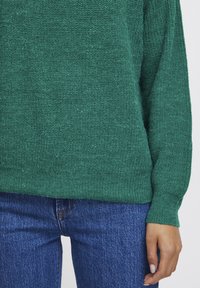 Groene gebreide trui met een losse pasvorm, geribde manchetten en een gestructureerde stof, gecombineerd met blauwe denim jeans.