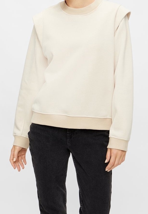 Personne portant un sweat-shirt beige à manches longues avec épaules rembourrées et un pantalon noir, vue du cou jusqu'aux hanches.