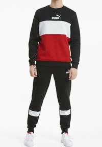 Felpa Puma nera e rossa con striscia bianca sul petto; pantaloni della tuta neri con strisce bianche alle caviglie; abbigliamento sportivo casual.