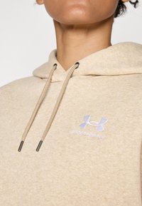Beige hoodie i mjukt tyg med dragsko. Har en ljuslila Under Armour-logga broderad på framsidan.