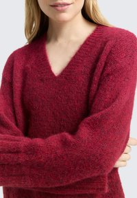 Głęboko czerwony sweter z dekoltem w serek wykonany z teksturowanej dzianiny. Posiada długie, luźne rękawy i luźny krój. Całość ma miękki, puszysty wygląd.