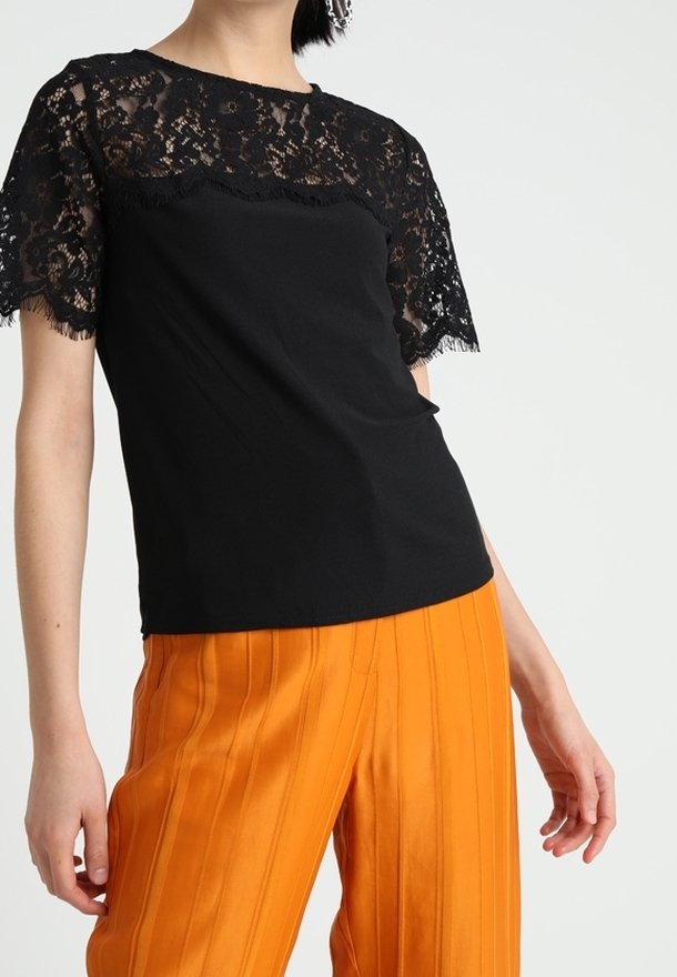 Top negro con mangas cortas de encaje y escote redondo, combinado con pantalones naranjas brillantes plisados y entallados. Textura de tela suave, colores contrastantes.
