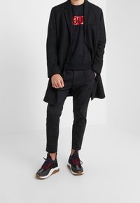 Blazer negro sobre una sudadera negra con logo rojo, combinado con pantalones negros y zapatillas negras con cordones rojos y suela marrón.