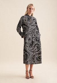 Schwarz-weißes Kleid mit Paisleymuster, langen Ärmeln, Taschen und hohem Kragen. Der Stoff hat eine glatte Textur.