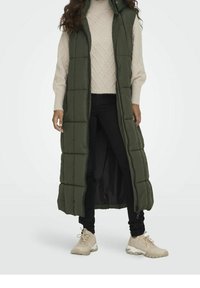 Person wearing a long olive green quilted sleeveless coat, cream knit sweater, black pants, and beige sneakers standing on a white background.