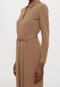 Femme portant une robe beige à manches longues avec un col et une ceinture assortie nouée à la taille, debout devant un fond blanc.
