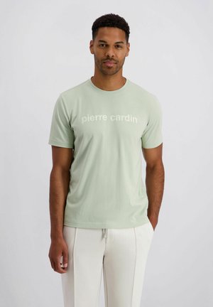 Man die vooruit staat, draagt een lichtgroen T-shirt met 'pierre cardin' en witte broek, handen ontspannen langs zijn zij tegen een effen achtergrond.