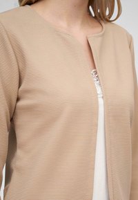 Beige žebrovaný kardigan s kulatým výstřihem a otevřeným předním dílem. Texturovaná tkanina. Nosený přes bílý top s krajkovými detaily na výstřihu.