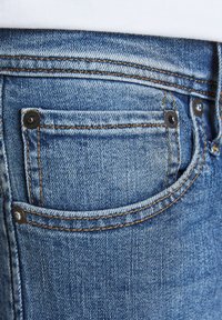 Τζιν παντελόνι σε μπλε απόχρωση, με μικρή πίσω τσέπη, ορατές ραφές σε αντίθετο πορτοκαλί χρώμα και μεταλλικά σκουλαρίκια.