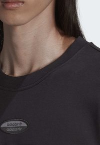 Čierna mikina s rebrovaným golierom, s textúrovaným oválnym logom adidas na hrudi. Hladká tkanina, priliehavý strih.