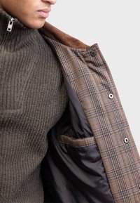 Homme portant un pull en maille côtelée vert foncé avec une demi-fermeture éclair, tenant ouverte une veste à carreaux marron montrant sa doublure intérieure noire et sa poche.