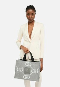 Donna in un completo crema tiene in mano una borsa a tracolla grigia con manici neri su uno sfondo bianco.