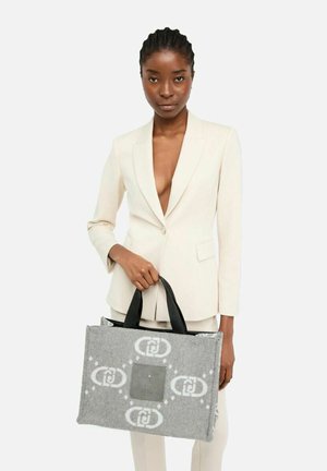 Mujer con un traje crema sosteniendo un bolso tote gris con un patrón y asas negras, contra un fondo blanco.