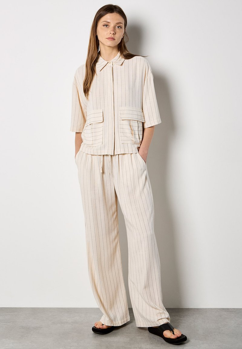 Jeune femme portant un pantalon ample crème à fines rayures et une chemise à manches courtes avec de grandes poches avant, debout contre un mur blanc.