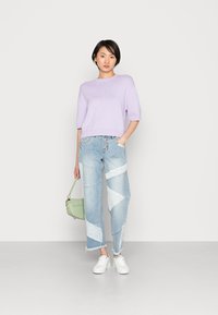 Suéter de malha lilás claro com mangas curtas, calças de ganga desgastadas com um design de patchwork geométrico e ténis brancos. Bolsa verde.