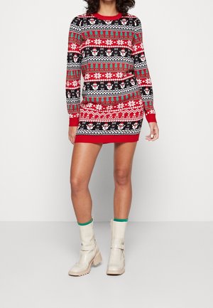 Vrouw draagt een feestelijke kersttrui-jurk in rood, wit, zwart en groen met sneeuwpoppen en boom patronen, gecombineerd met crèmekleurige enkellaarsjes.