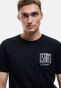 Czarny bawełniany T-shirt z okrągłym dekoltem. Z przodu na lewej piersi widnieje biały napis graficzny "STAY Classic". Krótkie rękawy.