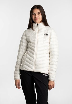 Doudoune blanche isolée avec un design matelassé vertical, col haut et fermeture éclair à l'avant. Présente un petit logo noir sur la poitrine.