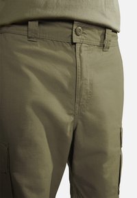 Pantaloni cargo in tessuto verde oliva, con chiusura a bottone, fronte piatto e tasche laterali con cuciture visibili e una texture liscia.