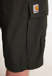 Shorts cargo vert foncé avec une poche à rabat et un logo Carhartt visible sur le côté, portés par une personne à la peau claire visible sous l'ourlet.