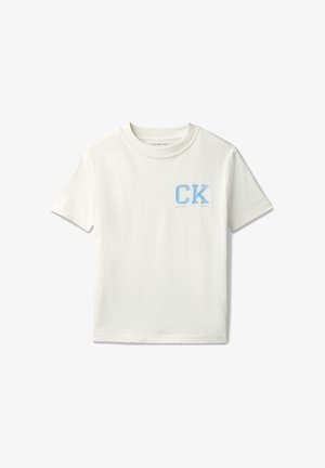 Jongen met lichtblauwe emmerhoed, off-white T-shirt met "CK"-logo, lichtblauwe spijkerbroek en witte sneakers, staand met handen in zijn zakken.