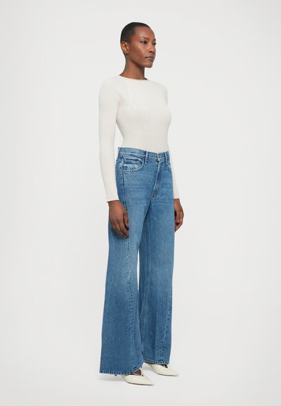 Light beige ribbed long-sleeve top paired with high-waisted, wide-leg blue denim jeans. White pointed-toe heels complete the outfit.